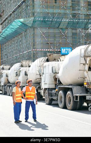 Asian male construction workers looking away discussing project while standing together near cement mixer trucks in construction site Stock Photo