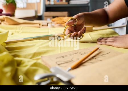 Cropped image of Black female fashion designer sitting cutting yellow fabric using scissors on table with ruler, pencil, and fashion sketchbook in ... Stock Photo