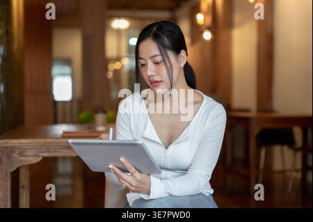 Asian woman using stylus pen and tablet while sitting with crossed legs on chair in coffee shop Stock Photo