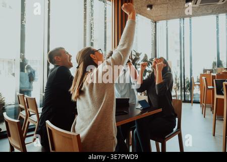 Cheerful Diverse group of business people celebrating with arms raised while sitting at table in cafe Stock Photo