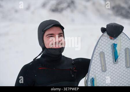 Smiling Mixed race man in surfing suit holding surf board and looking away while standing on snowy mountain during winter Stock Photo