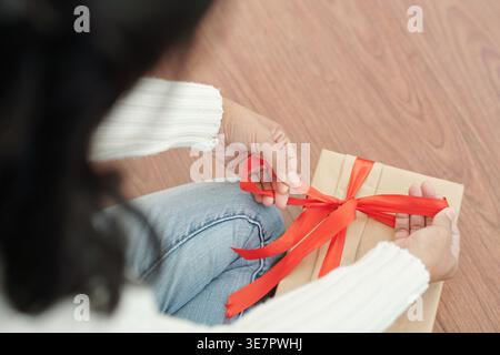 Cropped image of Vietnamese woman in white sweater tying red ribbon on Christmas present while sitting on floor at home Stock Photo
