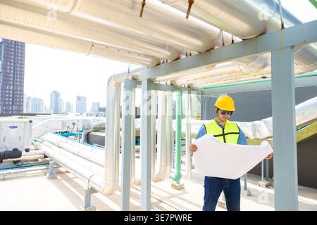 Asian male engineer wearing safety helmet checking blueprints standing near pipeline on rooftop during daytime Stock Photo
