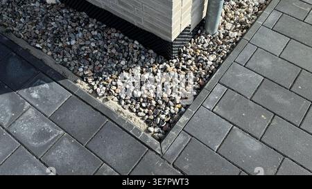 Close-up view of a corner of a paved patio featuring gray interlocking stones and decorative gravel surrounding a brick column with a drainage pipe Stock Photo
