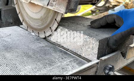 Worker using a concrete saw to cut a block of stone on a cutting table outdoors, with a blurred background of construction materials and tools Stock Photo