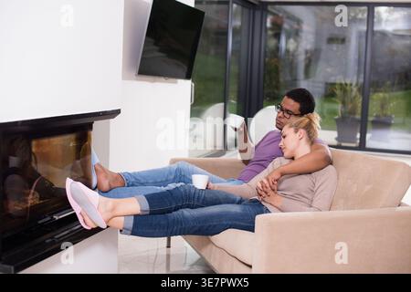 Middle Eastern man embracing caucasian wife while having coffee with their feet placed near fireplace sitting close together on sofa at home Stock Photo
