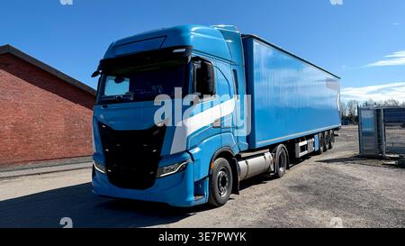 Blue heavy-duty truck with a large trailer parked on a gravel surface beside a red brick building under a clear blue sky Stock Photo