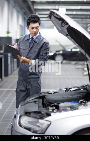 Serious Chinese male mechanic standing with clipboard checking car engine in repair shop Stock Photo