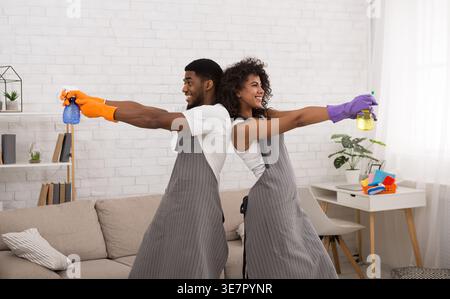 Smiling Black couple with aprons looking away holding spray bottles standing back to back in apartment Stock Photo