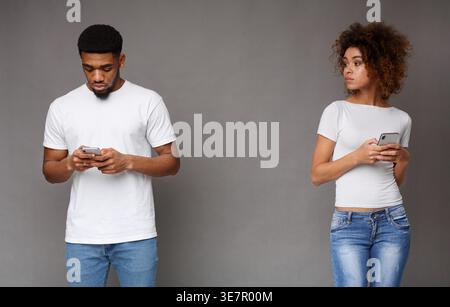 Suspicious Black woman looking at her focused Black man partner texting against gray background in studio Stock Photo