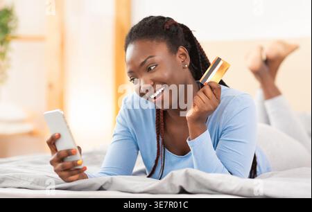 Smiling Black woman with braids holding credit card while using smartphone lying down in bed at home Stock Photo
