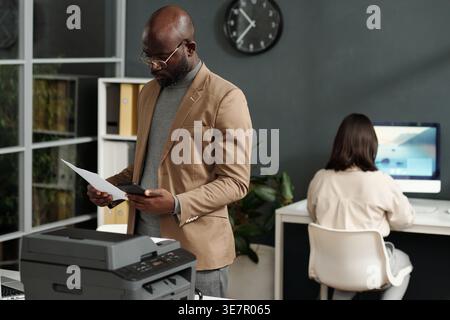 Black businessman looking at his smartphone together with documents standing nearby of printing machine in office Stock Photo