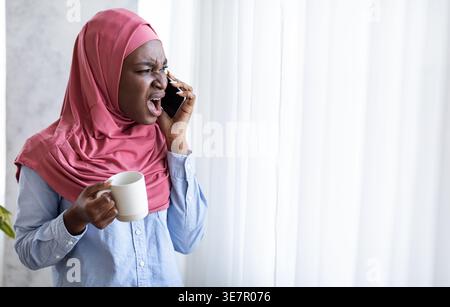 Angry Black woman wearing hijab holding cup of coffee having phone call while standing near window in living room at home Stock Photo