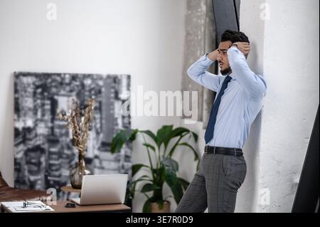 Arab businessman with sad expression and hands behind head looking away standing and leaning on wall near table with laptop at office Stock Photo