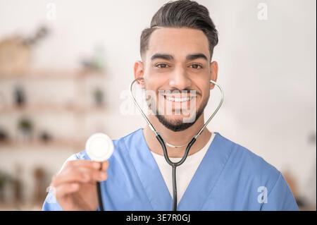 Smiling Arab male doctor holding stethoscope standing at home Stock Photo