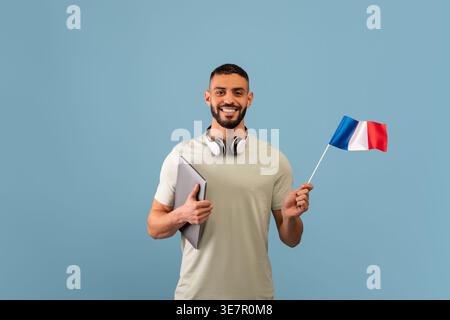Smiling Middle Eastern man with beard wearing headphones on neck holding laptop and flag of France standing against blue background in studio Stock Photo