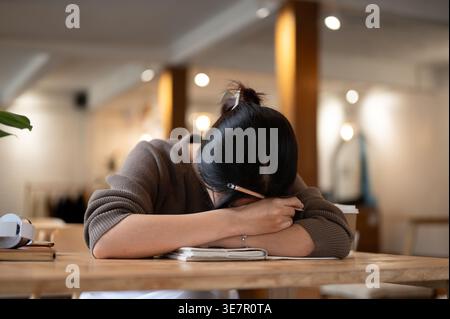 Asian woman holding pencil leaning on books while sitting and falling asleep at table in coffee shop Stock Photo