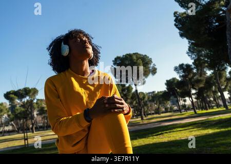 Black woman with headphones stretching legs in park on sunny day Stock Photo