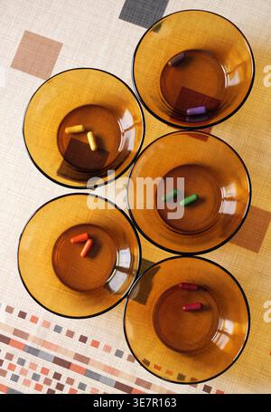 Five amber glass bowls with colorful dye capsules for Easter eggs arranged on patterned tablecloth, with visible shadows and warm indoor lighting 2026 Stock Photo