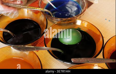 People wearing gloves dye Easter eggs using colorful liquids, with hands holding multicolored eggs and bowls of blue and dark dyes on a festive table Stock Photo