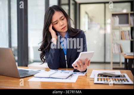 Stressed Asian business woman touching her temple and looking at calculator while sitting at her desk in the office Stock Photo