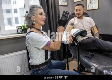 Smiling Asian male client sitting on chair and looking at middle-aged caucasian female tattoo artist putting on latex gloves preparing for tattoo s... Stock Photo