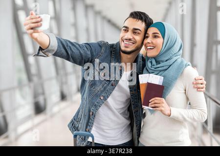 Middle Eastern couple taking selfie using smartphone and holding passport with ticket standing close together in airport Stock Photo