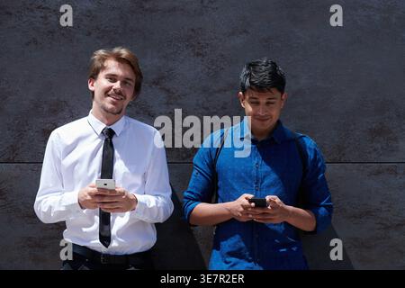 Middle Eastern and caucasian men using smartphones standing against concrete wall outdoors on sunny day Stock Photo
