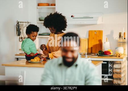 Smiling Black mother feeding her son sitting on kitchen counter near Black father working remotely on breakfast table at home Stock Photo