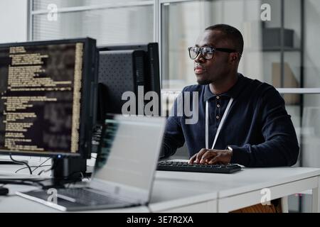 Focused Black male programmer in eyeglasses sitting at desk typing computer codes during work in office Stock Photo