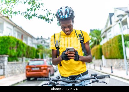 Black man wearing helmet and delivery bag using smLifestylephone to contact customer while standing with bicycle on street during daytime Stock Photo