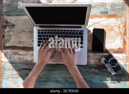 Black woman using laptop sitting at table with camera and smartphone at home on sunny day Stock Photo
