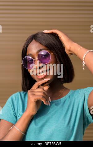 Smiling Black female in sunglasses touching hair and face while standing against brown wall Stock Photo