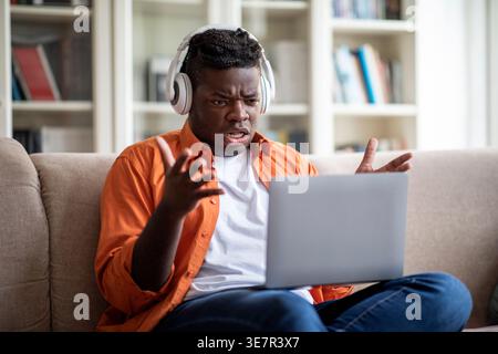 Upset Black man wearing headphones having online meeting sitting on sofa in living room at home Stock Photo