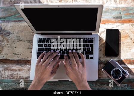 Black woman using laptop sitting at table with camera and smartphone at home on sunny day Stock Photo