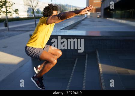 Black man with afro wearing yellow shirt looking away while doing squats with jumping on stairs in city on sunny day Stock Photo