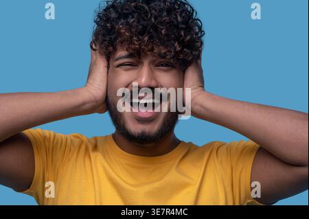 Indian man with curly hair standing with hands covering ears and mouth opened against blue background Stock Photo
