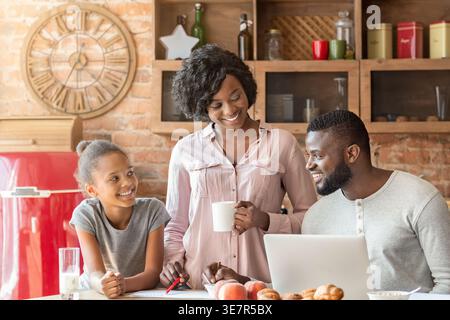 Smiling Black father working on laptop sitting at table near wife and daughter drinking milk and coffee looking at him having breakfast in kitchen ... Stock Photo