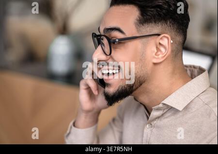Smiling Arab man wearing eyeglasses having phone conversation sitting at home Stock Photo