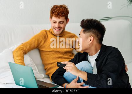 Cheerful Hispanic and caucasian gay couple purchasing online using credit card and laptop while sitting on sofa at home Stock Photo