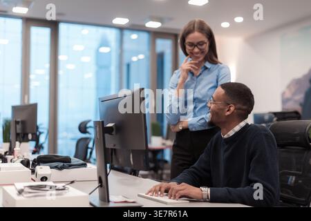 Black male working on computer sitting at desk talking to caucasian female coworker standing beside him in office Stock Photo