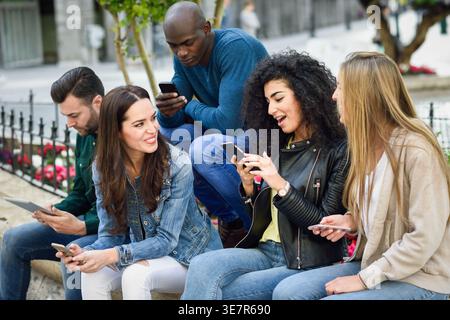 Smiling diverse group of friends using smartphones and having conversation while sitting together on bench in city park Stock Photo