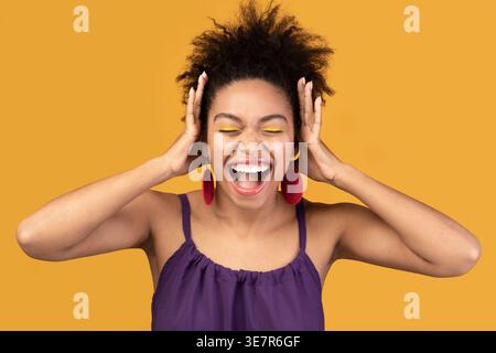 Black woman with closed eyes and open mouth screaming with hands on head standing against yellow background in studio Stock Photo