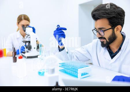Indian male scientist working with chemical liquids in flask near caucasian female colleague using microscope in laboratory Stock Photo