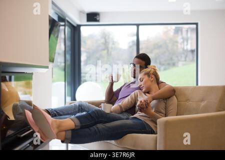 Middle Eastern man embracing caucasian wife while having coffee with their feet placed near fireplace sitting close together on sofa at home Stock Photo