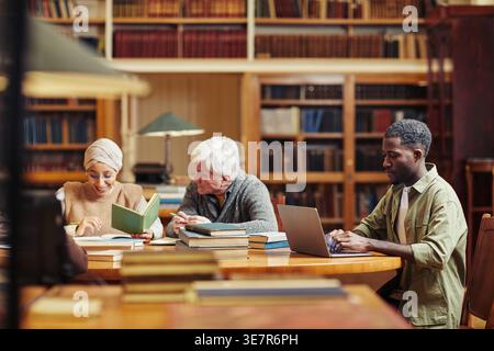 Serious Black man using laptop sitting at table near diverse friends with books in library Stock Photo