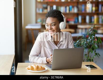 Black woman with afro wearing headphones listening to music looking away sitting in front of laptop at table in coffee shop Stock Photo