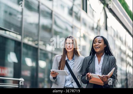 Smiling Black businesswoman with braids holding book talking to caucasian female colleague with tablet walking outside office building during daytime Stock Photo