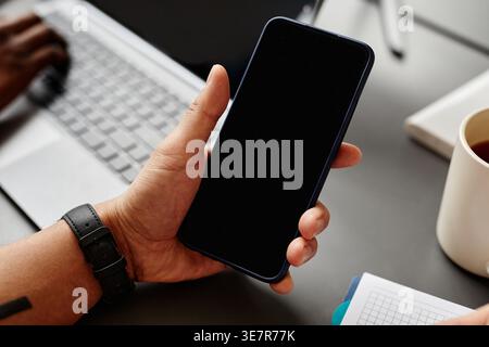Hand of Asian male programmer holding smartphone with codes on screen sitting at table with laptop in office Stock Photo