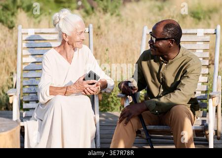 Middle-aged Black man with crane and senior caucasian woman looking at each other sitting on chair in park on sunny day Stock Photo